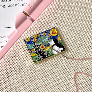 Könnte beinhalten: Ein rosa Stickrahmen mit aufgespanntem Stoff. Eine kleine Emaille-Anstecknadel mit Van Goghs "Sternennacht" und einer Katze ist auf die Leinwand gesteckt. Eine Nadel und rosa Faden sind ebenfalls zu sehen.