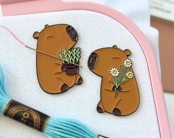May include: Two brown capybara enamel pins attached to white fabric in a pink Nurge embroidery hoop. One pin shows a capybara holding a potted plant, the other a capybara holding a bouquet of daisies. Two skeins of blue embroidery thread are included.
