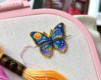May include: A light pink Nurge embroidery hoop holding white fabric with a butterfly-shaped needle minder featuring a blue, yellow, and purple starry night pattern. A silver needle is attached to the minder, with orange and pink embroidery floss.