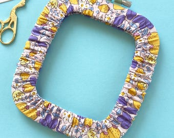 May include: A square embroidery hoop cover featuring a light pink fabric with a repeating pattern of yellow strawberries, purple flowers, and green leaves. The cover includes a green tension screw and is accompanied by gold scissors and three skeins of embroidery floss in light blue, purple, and yellow.