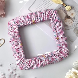 May include: A square, floral-patterned fabric frame cover in pink, white, and blue. The cover is ruffled along the edges. A spool of yellow thread and a strand of pink pearls are also visible.