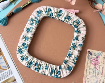 May include: A square embroidery hoop cover made of cream-colored fabric with a repeating pattern of small blue roses and green leaves. The cover features a gathered, elasticized edge designed to fit snugly around the frame of the hoop.