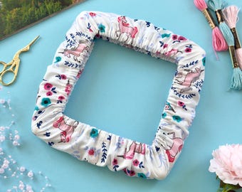 May include: A square fabric embroidery hoop cover featuring a white background with a repeating pattern of pink unicorns, blue and pink flowers, and green leaves. The cover has an elasticized edge and is gathered around the square frame.