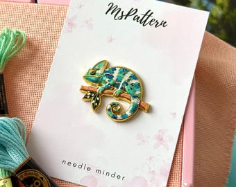 May include: A gold-toned enamel needle minder featuring a chameleon perched on a branch, displayed on a white card with the text MsPattern and needle minder. The chameleon has blue, green, and white patterned scales with gold outlines.