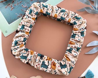 May include: A square fabric embroidery hoop cover featuring a repeating pattern of various cats in shades of blue, orange, and brown on a cream background. The cover has an elasticized edge to fit securely around a square frame for needlework projects.