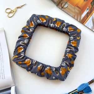 May include: A square fabric embroidery hoop cover in a dark grey colour featuring a repeating pattern of orange foxes, rainbows, stars, and moons. The cover has an elasticated edge to fit securely around a square frame for needlework projects.