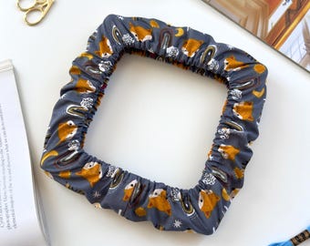 May include: A square fabric embroidery hoop cover in a dark gray color featuring a repeating pattern of orange foxes, rainbows, stars, and moons. The cover has an elasticized edge to fit securely around a square frame for needlework projects.