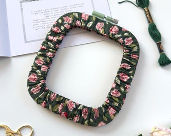May include: A square embroidery hoop wrapped in dark green fabric featuring a pink rose floral pattern. The hoop includes a light green tightening screw at the top. The design is characterized by gathered fabric texture around the frame.