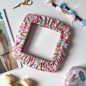 May include: A square fabric frame cover featuring a pink, blue, and green floral pattern with gathered edges. The item is displayed alongside gold embroidery scissors, two skeins of embroidery floss, a magazine, a heart-shaped tin, and tulips.