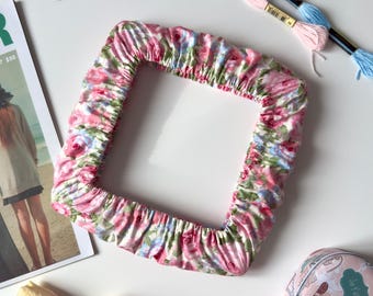 May include: A square fabric frame cover featuring a pink, blue, and green floral pattern with gathered edges. The item is displayed alongside gold embroidery scissors, two skeins of embroidery floss, a magazine, a heart-shaped tin, and tulips.