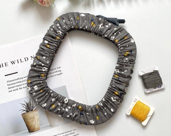 Puede incluir: Una diadema de tela gris con un estampado floral de flores blancas y amarillas. La diadema es circular y fruncida. Una revista, carretes de hilo y una ramita de flores también están en la imagen.
