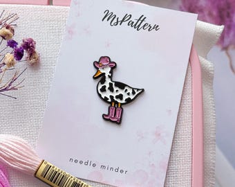 May include: A needle minder with a cartoon duck wearing a pink cowboy hat and boots, with black and white spots. The needle minder is on a card that says "MsPattern" and "needle minder". Pink and white embroidery floss is in the foreground.