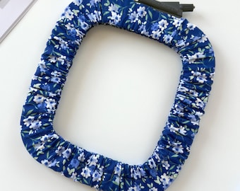 May include: A square embroidery hoop cover featuring a blue fabric with a white and light blue floral pattern. The cover is gathered with elastic to fit around the frame, with a black plastic tension screw visible at the top corner.
