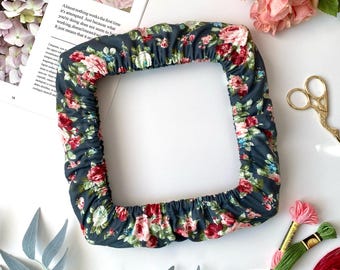 May include: A square, floral-patterned object with a dark teal background and red and white flowers. The object is surrounded by pink roses, embroidery floss, and a magazine with the text "WIND AGAIN."