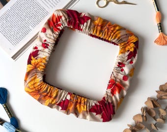 May include: A square fabric embroidery hoop cover featuring a pattern of red, orange, and yellow autumn maple leaves on a light background. The cover has an elasticized edge and is surrounded by embroidery floss, gold scissors, and a book.
