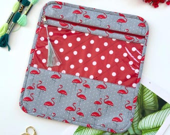 May include: A square quilted pouch featuring a gray fabric with a red flamingo and white polka dot pattern. It includes a clear vinyl pocket with red and white polka dots, a red zipper with a silver tassel pull, and rounded corners.