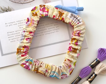May include: A square fabric embroidery hoop cover featuring a Rilakkuma character print with yellow, pink, and brown accents. The cover has an elasticized edge and a small tag labeled Rilakkuma. Two skeins of embroidery floss in purple and cream.