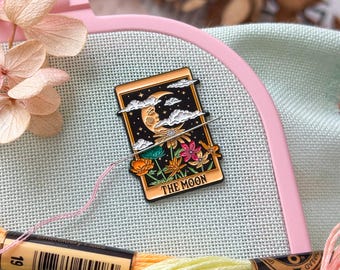 May include: An enamel pin featuring a tarot card design with a crescent moon, clouds, and flowers. The pin is set on a pink embroidery hoop with colorful embroidery floss in shades of orange, yellow, and pink. Dried flowers are also visible.