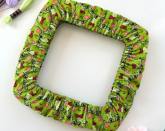 May include: A square fabric embroidery hoop cover featuring a repeating pattern of green frogs with pink cheeks and small red bows. The cover has an elasticized edge designed to fit snugly around a square frame, showcasing a vibrant cartoon print.