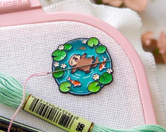 May include: A pink embroidery hoop holds white fabric with a colorful pin featuring an otter in water with fish and lily pads. Green embroidery floss and a needle are also visible. The hoop is labeled "Nurge".