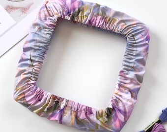 May include: A square embroidery hoop cover featuring a soft lavender and purple floral pattern with an elasticized edge. The fabric is gathered around the frame, creating a ruffled appearance. Three skeins of embroidery floss in blue and purple shades.