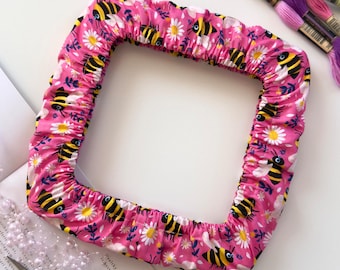 May include: A square fabric embroidery hoop cover in a vibrant pink color featuring a repeating pattern of yellow and black bees, white daisies, and small blue floral sprigs. The cover has an elasticized edge for a snug fit around the frame.