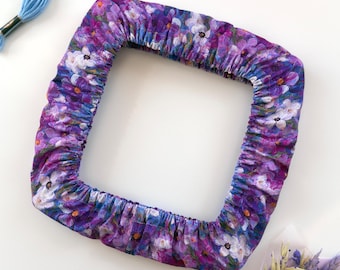 May include: A square fabric embroidery hoop cover featuring a vibrant purple, blue, and white floral pattern with gathered edges. Two skeins of embroidery floss in light purple and light blue, and a small bouquet of dried flowers are nearby.