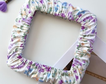 May include: A square embroidery hoop cover made of white fabric featuring a floral pattern in shades of purple, blue, and yellow. The cover has an elasticized edge for a snug fit. Four skeins of embroidery floss in blue, purple, and peach are included.