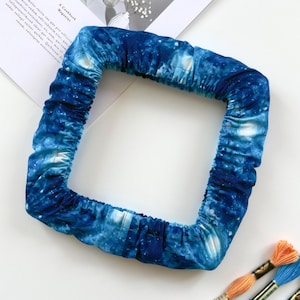 May include: A square embroidery hoop cover featuring a deep blue galaxy print with white nebula accents. The fabric is gathered with an elastic band to fit securely around the frame, creating a soft, textured border with a celestial pattern.