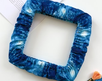 May include: A square embroidery hoop cover featuring a deep blue galaxy print with white nebula accents. The fabric is gathered with an elastic band to fit securely around the frame, creating a soft, textured border with a celestial pattern.