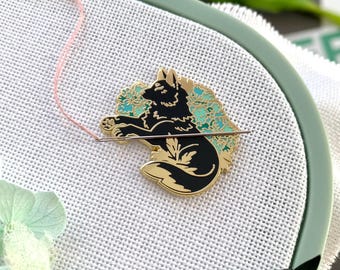 May include: A black and gold enamel pin with a wolf design, set against a teal and gold floral backdrop. The pin is on white fabric within a green embroidery hoop. Pink thread is also visible.