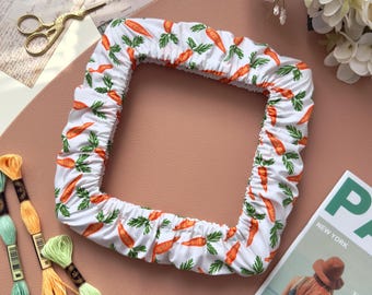 May include: A white fabric frame cover with a repeating pattern of orange carrots and green leaves. The cover is on a pink surface, with embroidery floss, scissors, and a magazine nearby.