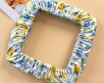 May include: A square embroidery hoop cover made of white fabric featuring a floral pattern with blue and yellow flowers. The cover has an elasticized edge that creates a gathered, ruffled effect around the square frame of the hoop.