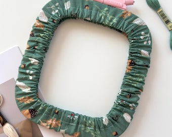 May include: A square embroidery hoop cover made of green fabric featuring a pattern of brown acorns and white leaves. The cover is elasticized and includes a small pink plastic tension adjustment knob. Two skeins of embroidery floss are visible.