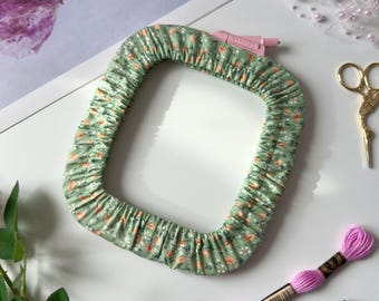 May include: A rectangular embroidery hoop cover in a light green fabric with a small floral pattern featuring orange and white flowers. The cover is gathered with elastic and includes a pink plastic tension adjustment knob at the top corner.