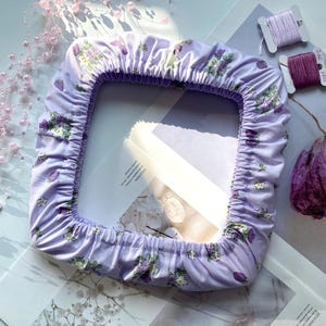 Puede incluir: Una funda cuadrada lavanda con borde elástico, adornada con un estampado floral de flores blancas y moradas y hojas verdes. La funda está sobre una superficie clara con elementos decorativos como cuentas y carretes de hilo.