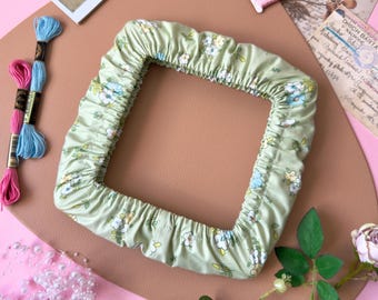May include: A square-shaped fabric frame cover in a light sage green color featuring a repeating pattern of small white, yellow, and blue flowers. The cover has an elasticized inner edge designed to fit snugly around a square embroidery hoop.