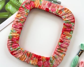 May include: A square embroidery hoop covered in a gathered, multi-colored floral fabric featuring shades of pink, orange, and green. The hoop includes a light green plastic tightening screw at the top. Three skeins of embroidery floss in mint green, light pink, and coral are positioned near the bottom right corner.