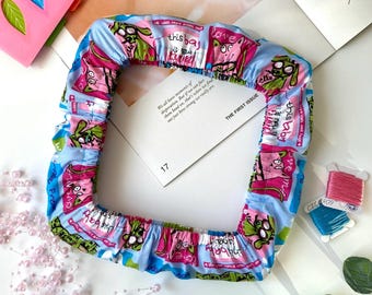 Puede incluir: Un artículo de tela cuadrado y con volantes con un estampado de dibujos animados en azul, rosa y verde. El texto "this baby" y "love" son visibles en la tela. El artículo está colocado sobre una superficie blanca con otros elementos decorativos.