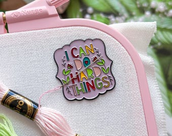 May include: A pink embroidery hoop holds white fabric with a colorful enamel pin that reads "I CAN DO HARD THINGS." Two skeins of embroidery floss, one pink and one green, are visible. The hoop is branded with "Nurge."