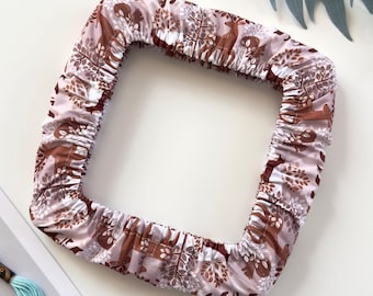 May include: A square fabric embroidery hoop cover featuring a repeating pattern of brown deer and stylized trees on a light pink background. The cover has an elasticized edge designed to fit snugly around the frame of a square embroidery hoop.