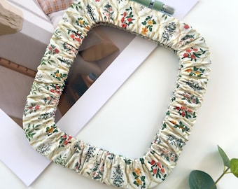 May include: A rectangular embroidery hoop cover made of cream-colored fabric featuring a repeating pattern of thin gold lines and small floral motifs in red, yellow, and green. The cover has an elasticized edge and a light green tension screw.