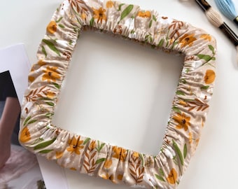 May include: A square fabric embroidery hoop cover featuring a beige background with a repeating pattern of yellow flowers, green leaves, and brown stems. The cover has an elasticized edge for a snug fit and is accompanied by two embroidery floss skeins.