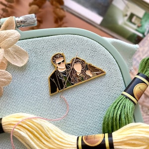 May include: A gold-colored enamel pin featuring a couple, with the man wearing sunglasses. The pin is set against a light blue embroidery hoop, with yellow and green embroidery floss and a needle. Dried flowers and a book are in the background.