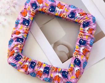 May include: A square fabric embroidery hoop cover in a light pink color featuring a repeating pattern of cartoon foxes, cats, and bananas. The cover has an elasticized edge for a snug fit and is shown with three skeins of embroidery floss.
