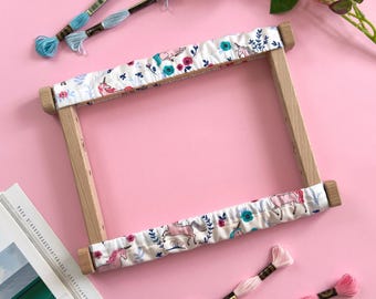 May include: A rectangular wooden embroidery frame with fabric covers featuring a pattern of pink unicorns, teal flowers, and blue leaves. Two light blue embroidery floss skeins and two pink embroidery floss skeins are placed nearby.