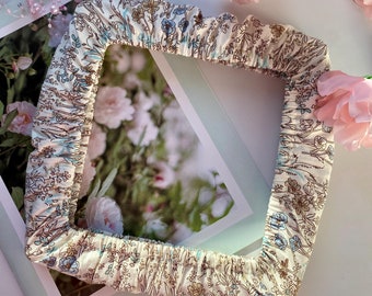 Puede incluir: Una diadema con estampado floral en forma cuadrada, con fondo crema y diseños florales azules y marrones. La diadema se coloca sobre una fotografía con flores rosas y un fondo claro.