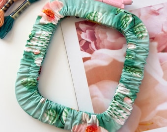 May include: A rectangular embroidery hoop cover made of light green fabric with a floral pattern featuring pink and white flowers and green leaves. The cover has an elasticized edge and a small pink plastic tension adjustment knob at the top.