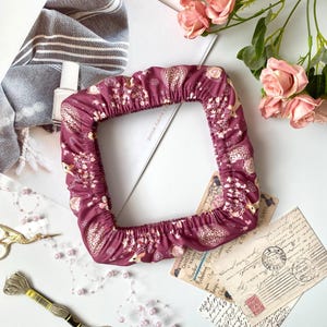 May include: A burgundy, square fabric cover with a repeating floral and animal pattern. The cover is gathered at the edges. The image includes pink roses, vintage postcards, and sewing supplies.
