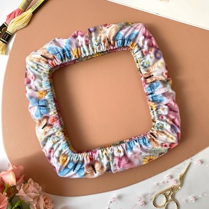 以下が含まれることがあります： ピンク、青、黄色の蝶が描かれた正方形のフローラル柄のアイテム。 桃色の表面に置かれ、刺繍糸、ハサミ、花が背景にあります。 手芸用品です。